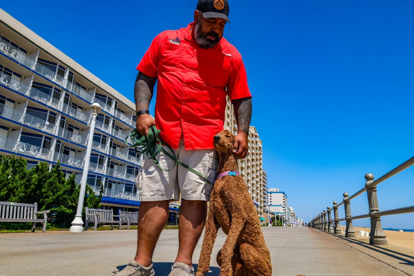 Trainer in a red shirt holding a green leash while a curly brown dog sits beside him on the Virginia Beach oceanfront boardwalk